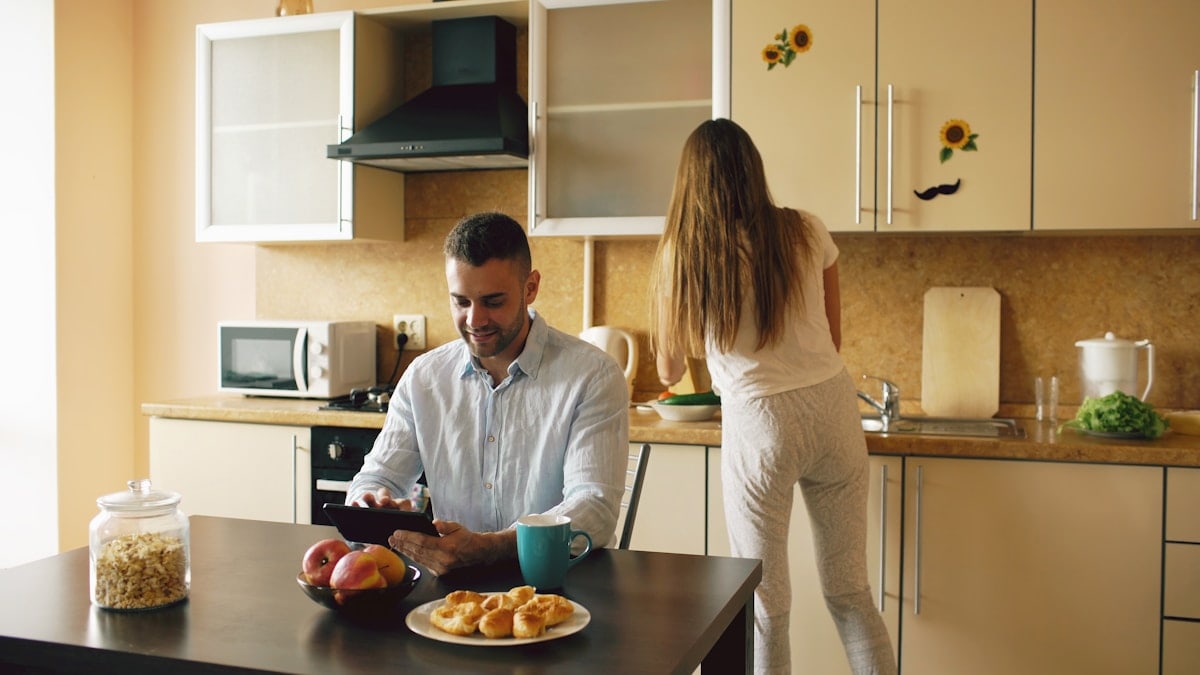 Happy young man using digital tablet computer sitting in the kitchen and talking his wife while she cooking at home in the morning